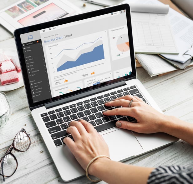Top-down view of a wooden table with laptops, documents and tablets, surrounded by illustrated gears and graphs, overlaid with the word ‘BUSINESS’ — conveying a corporate development and commercial growth concept.